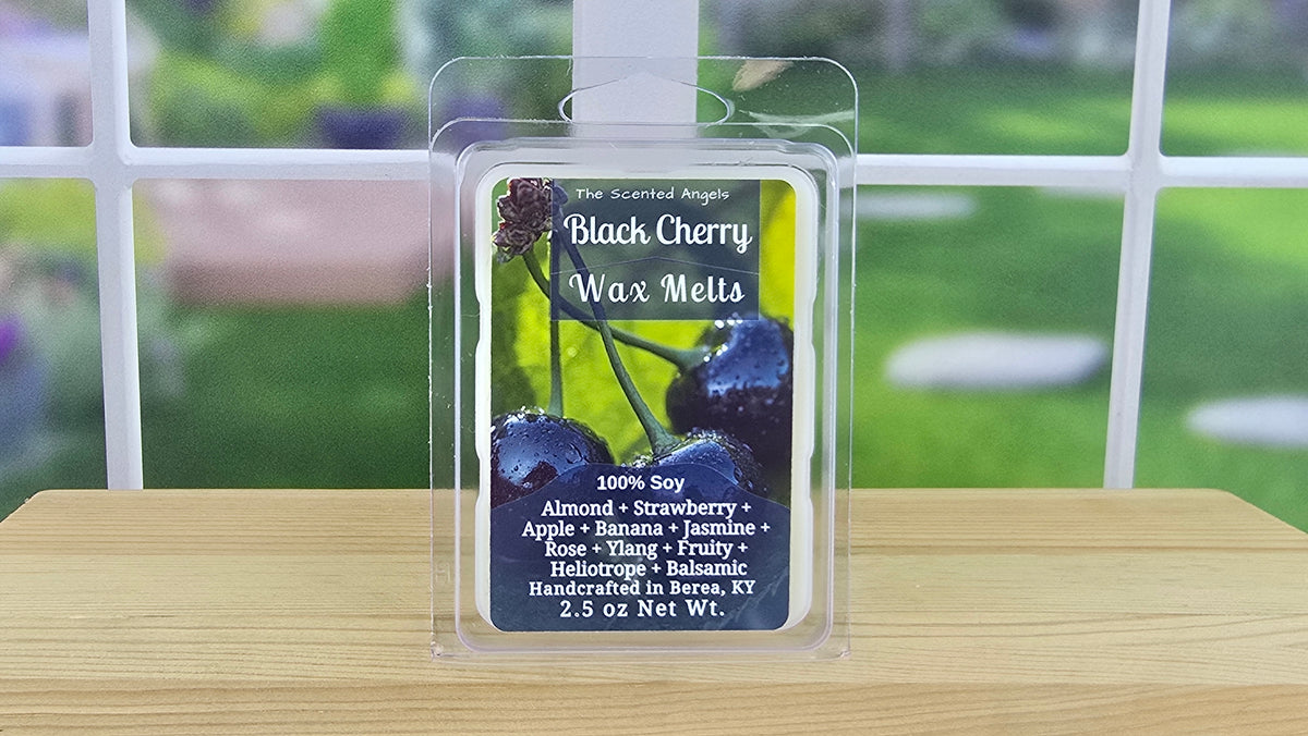 A packaged wax melt labeled 'Black Cherry Wax Melts' by The Scented Angel, shown in a clear plastic clamshell package on a wooden surface with a blurred window and green outdoor view in the background.