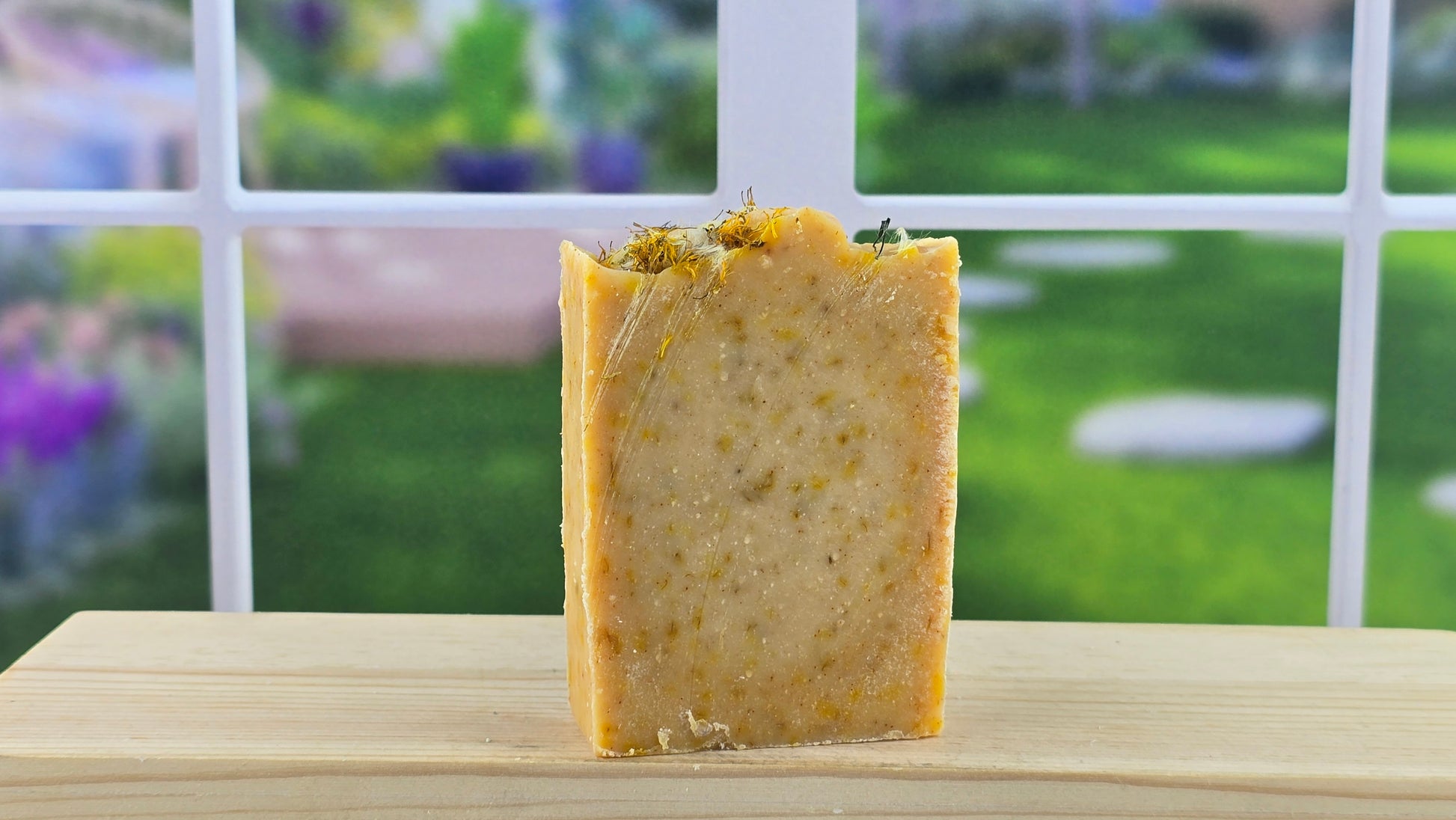 A bar of handmade soap, sprinkled with dandelion flowers, placed on a wooden surface with a bright window in the background.