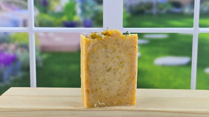 A bar of handmade soap, sprinkled with dandelion flowers, placed on a wooden surface with a bright window in the background.