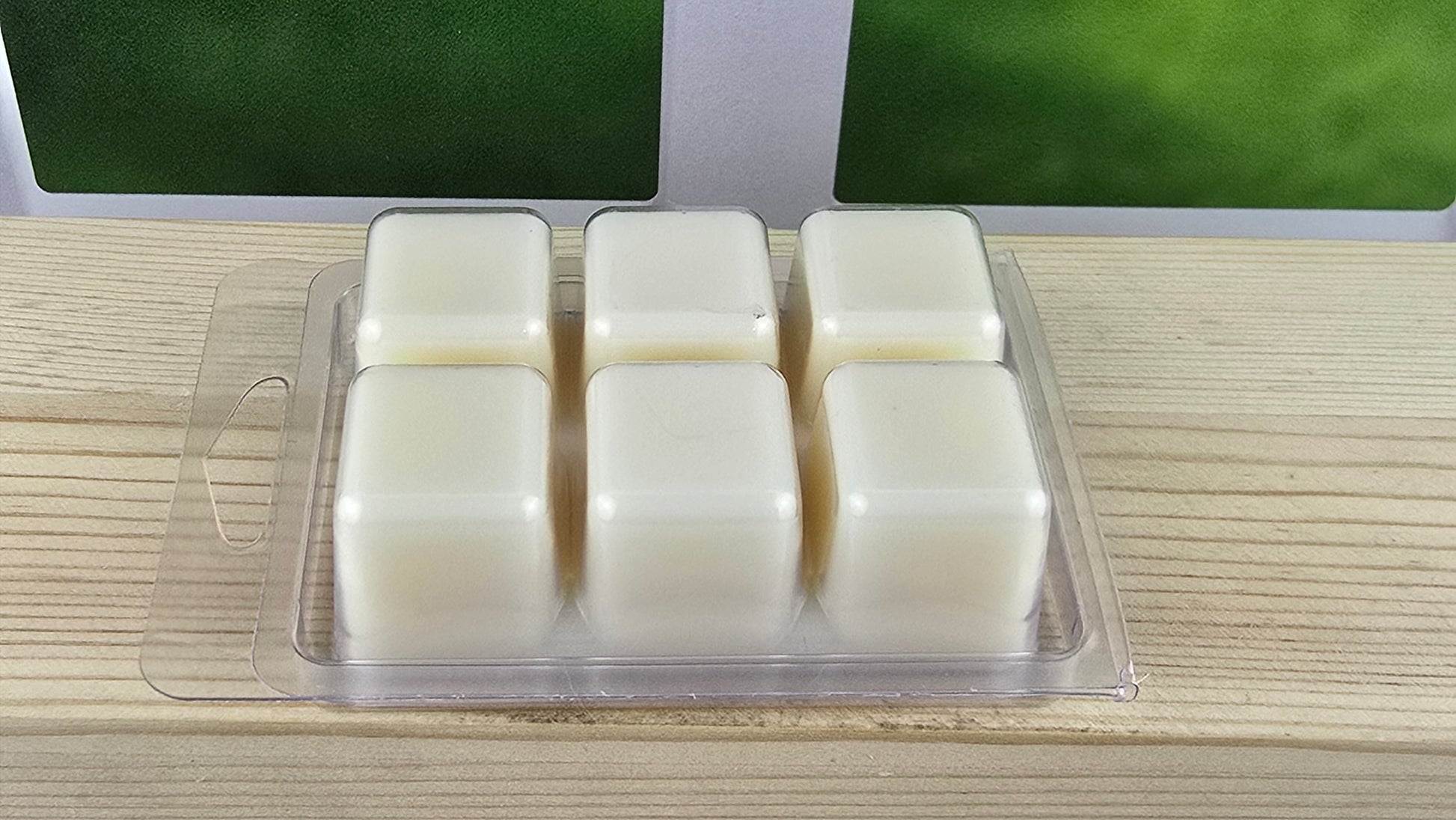 Six square wax melts in a clear plastic packaging on a wooden surface with a green grass background.