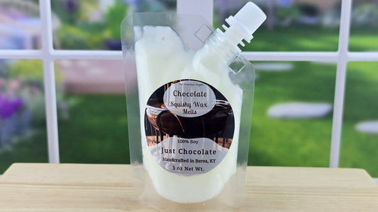 A clear squeezable wax melt pack labeled 'Chocolate' with a white cap, placed on a wooden surface with a bright background.