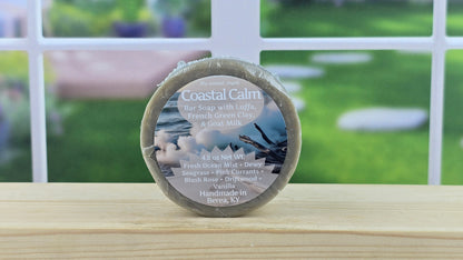 Coastal Calm bar soap with Luffa, French Green Clay and Goat Milk on a wooden surface with a blurred outdoor background.