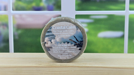Coastal Calm bar soap with Luffa, French Green Clay and Goat Milk on a wooden surface with a blurred outdoor background.