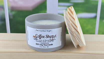 A white soy wax candle with a label reading 'Coffee Shop', set on a wooden surface with a bright background.
