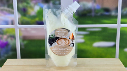 A pouch of Coffee Shop 100% Soy Squeezy Wax Melts placed on a wooden surface with a window in the background showing a grassy area.