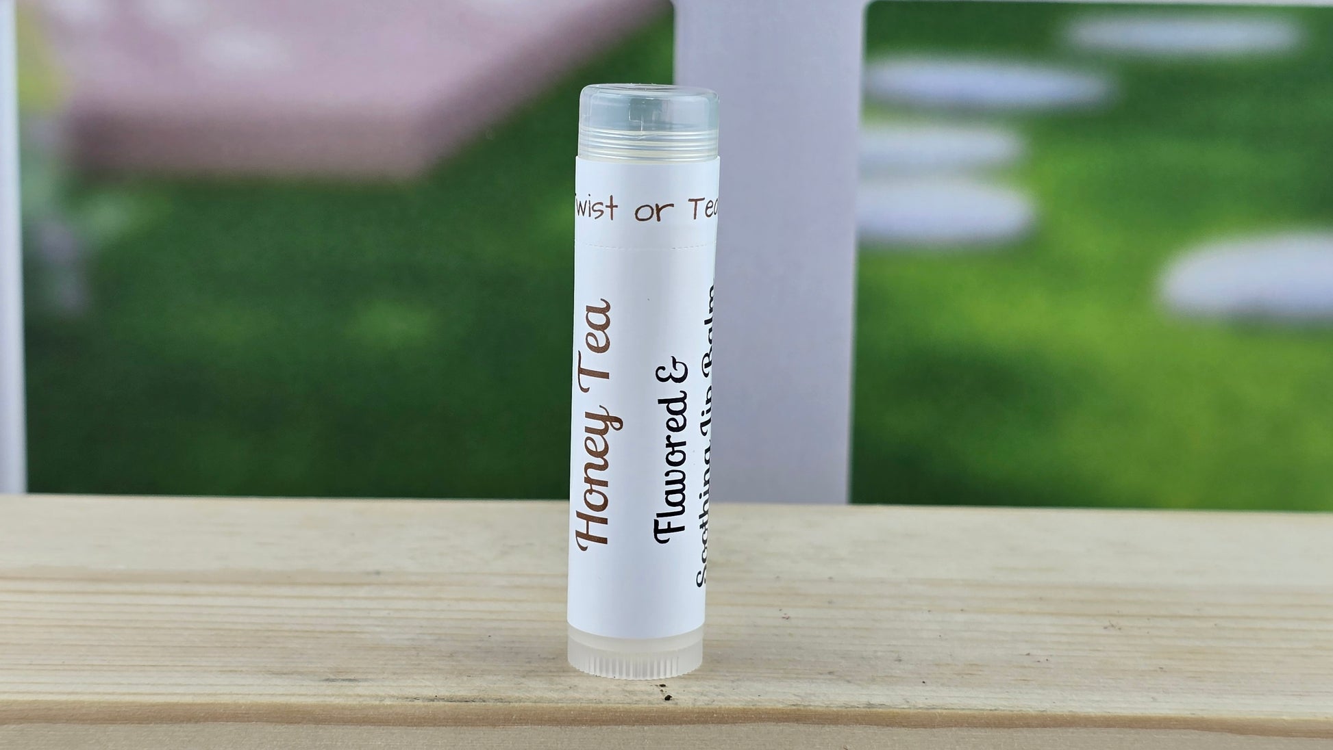 Clear lip balm tube labeled 'Honey Tea' on a wooden surface with a blurred outdoor background.