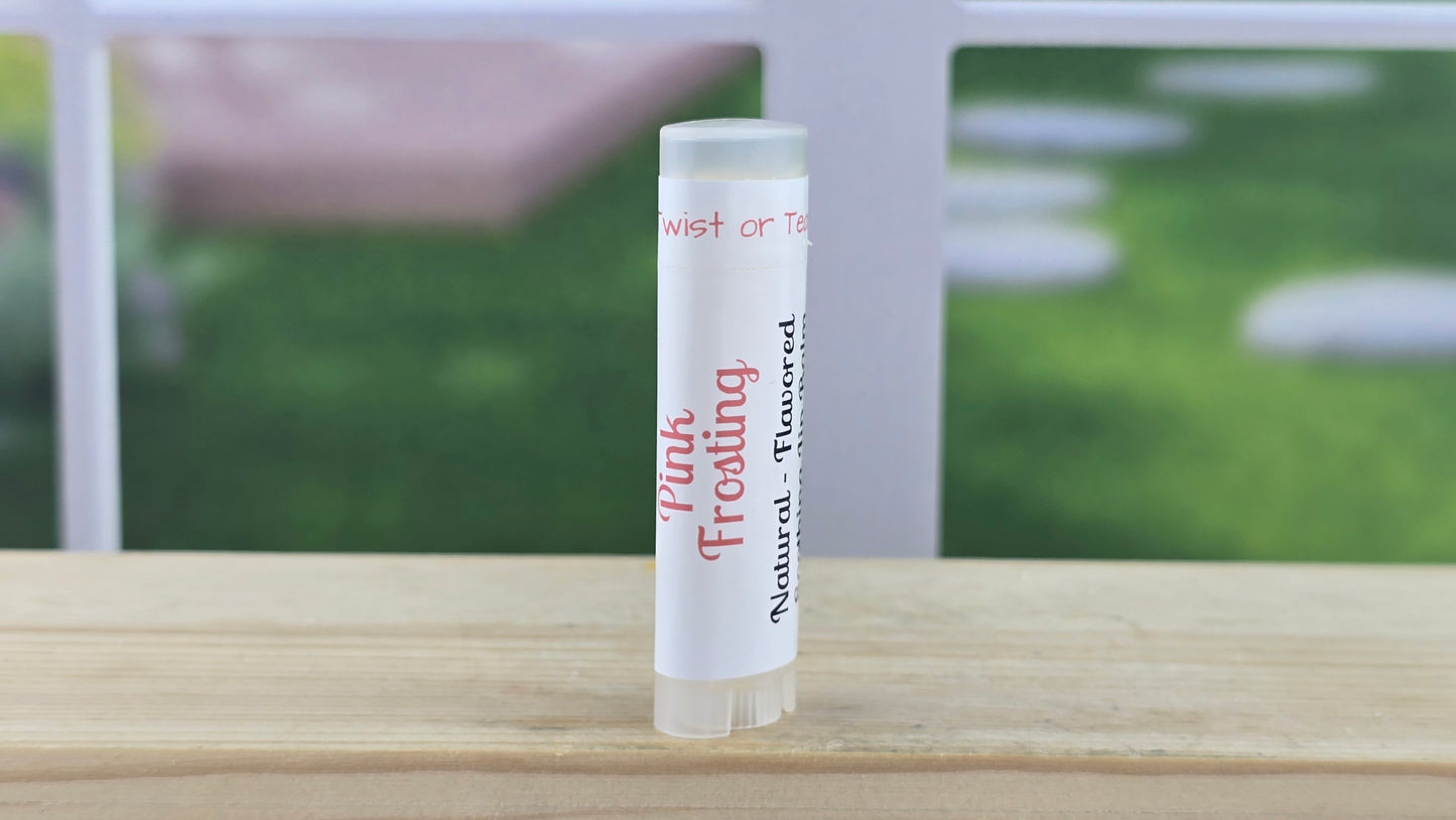 Lip balm tube labeled 'Pink Frosting' on a wooden surface with a blurred outdoor background