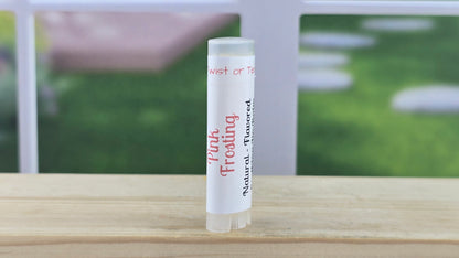Lip balm tube labeled 'Pink Frosting' on a wooden surface with a blurred outdoor background