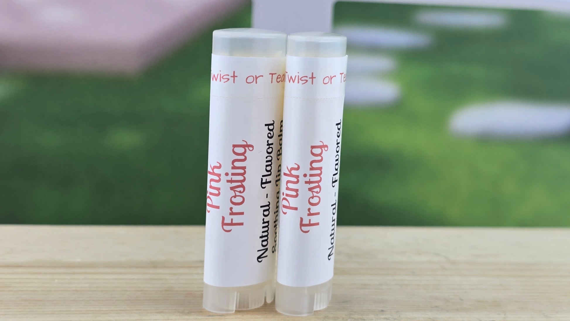 Pink Frosting lip balms on a wooden surface with a blurred green background.