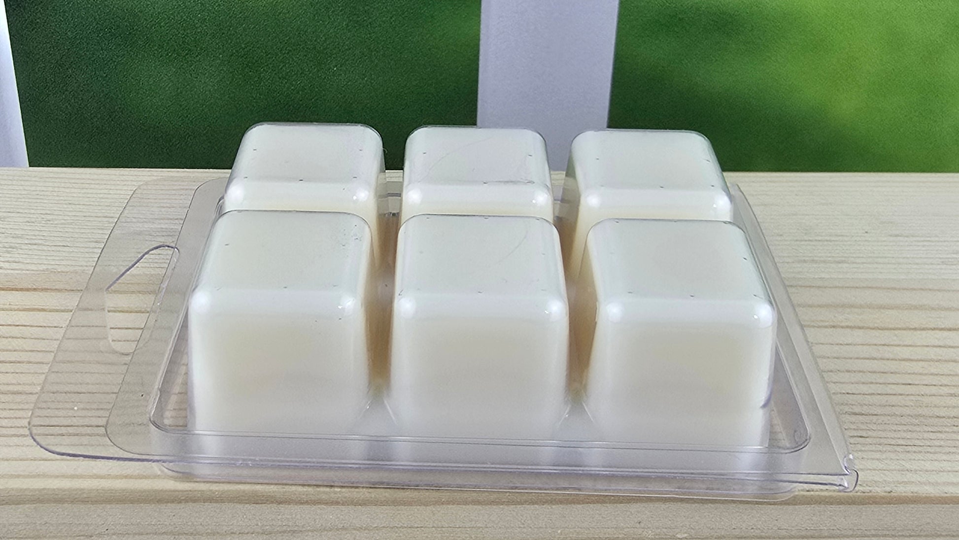 Set of six white wax melts in a clear plastic packaging on a wooden surface with a blurred green background.