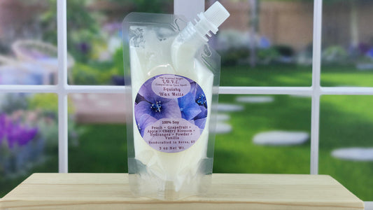A pouch of Love Spell 100% Soy Squeezy Wax Melts placed on a wooden shelf with a window in the background showing a garden and a floral display.