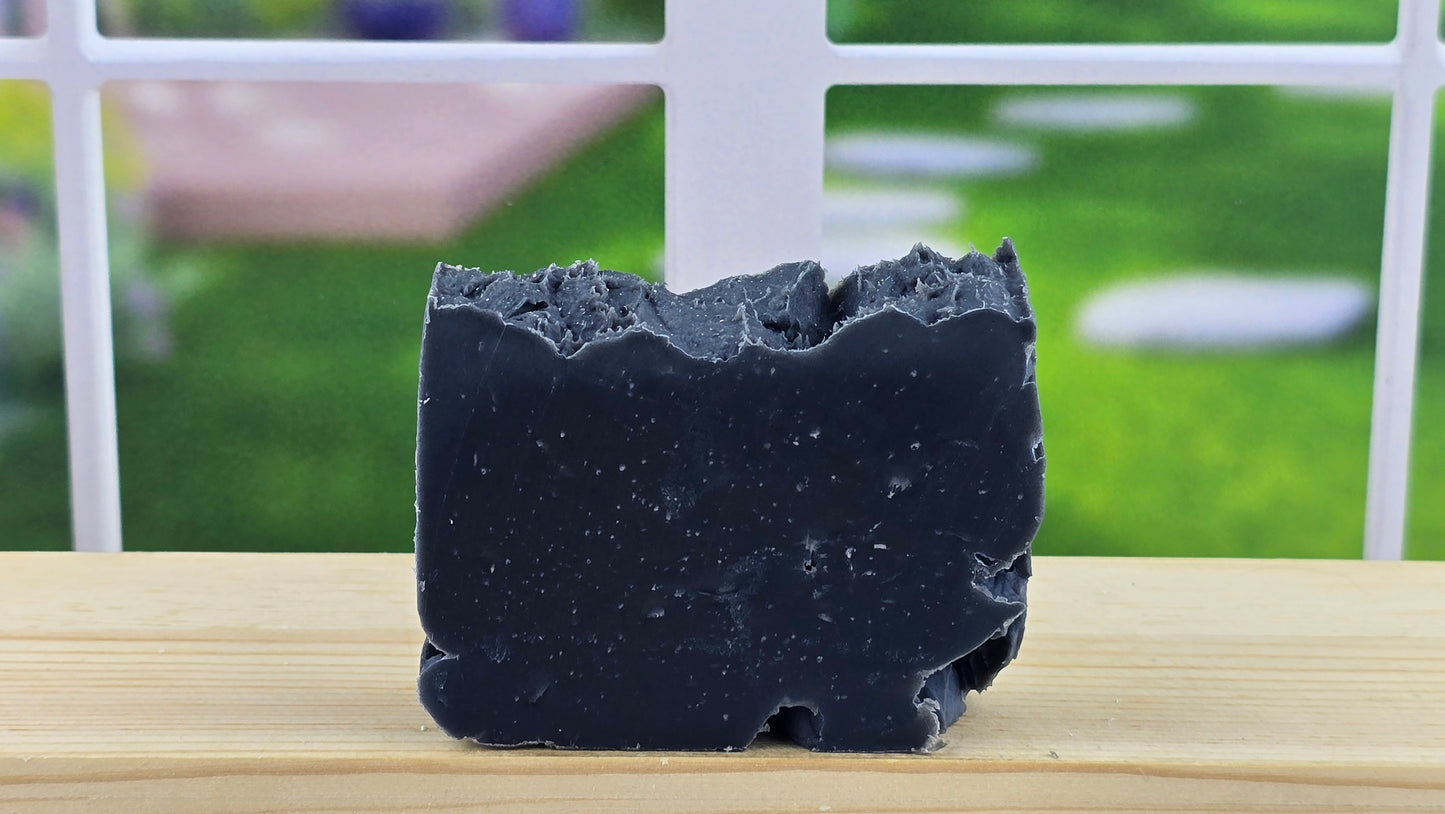 A bar of handmade Lump o' Coal cold process soap displayed on a wooden surface with a bright window in the background.