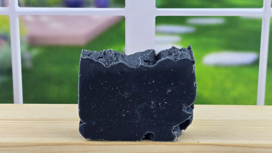 A bar of handmade Lump o' Coal cold process soap displayed on a wooden surface with a bright window in the background.