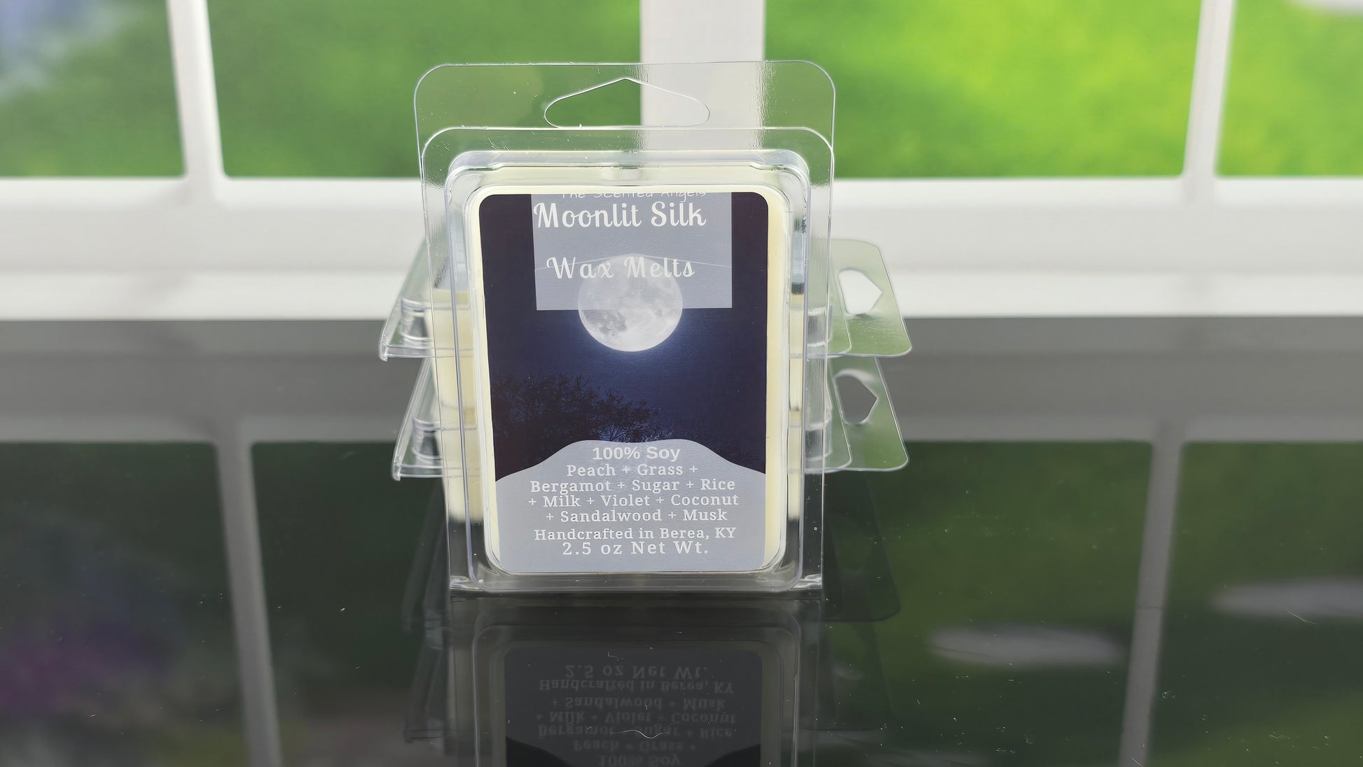 Packaged Moonlit Silk Wax Melts on a clear stand with a blurred green outdoor background