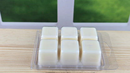 Four white wax melts in a clear plastic packaging on a wooden surface with a blurred green outdoor background.