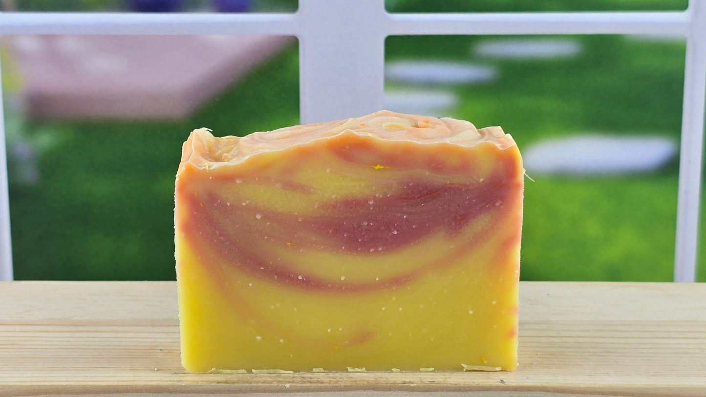 A bar of handmade Peach Sunset Cold Process Soap set on a wooden surface with a bright window in the background.