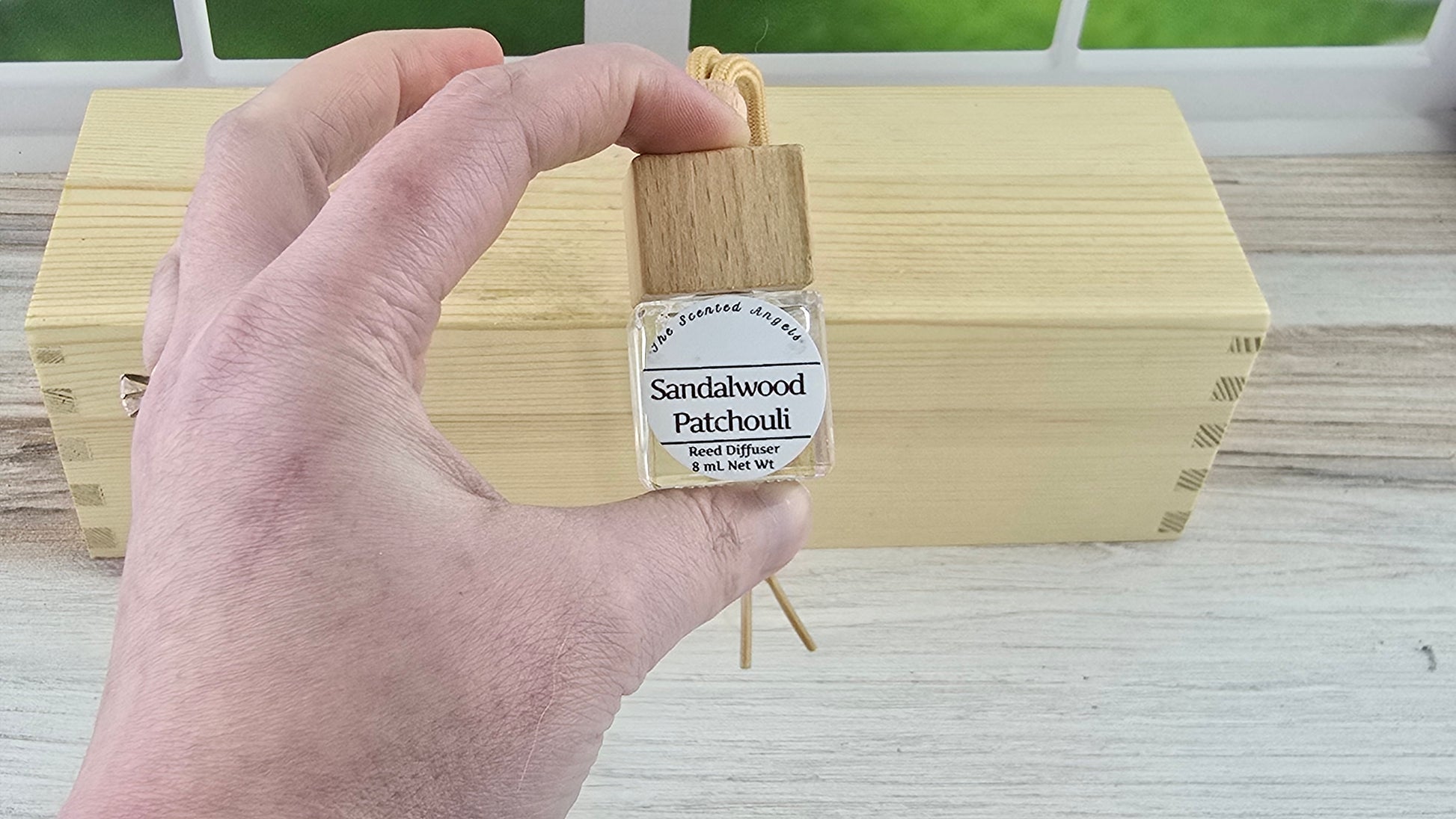 Hand holding a small bottle labeled 'Sandalwood Patchouli' in front of a wooden block.