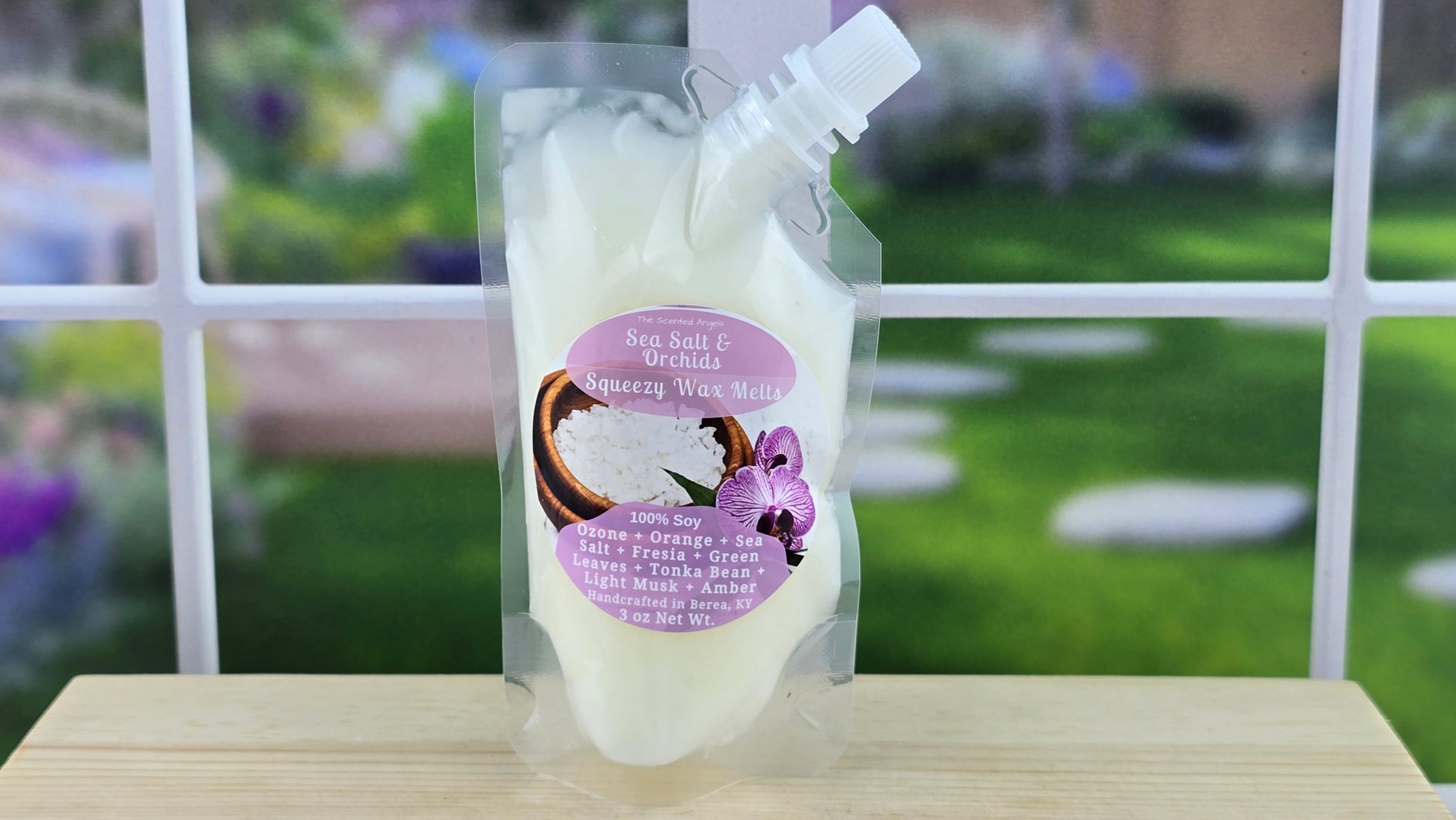 A clear plastic pouch filled with white wax melts, labeled 'Sea Salt & Orchids', placed on a wooden surface with a green, floral background.