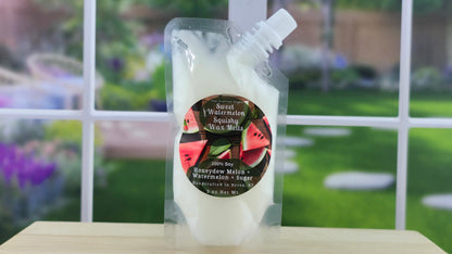 A clear plastic pouch containing white squeezable wax melts with a watermelon label on the front, placed on a wooden surface with a blurred greenery background.