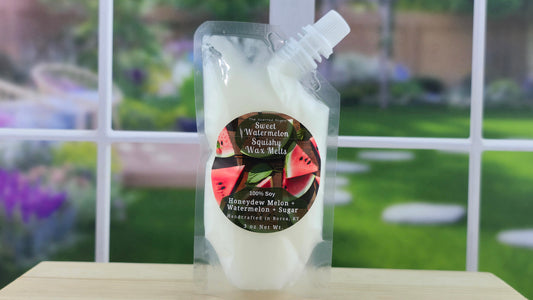 A clear plastic pouch containing white squeezable wax melts with a watermelon label on the front, placed on a wooden surface with a blurred greenery background.
