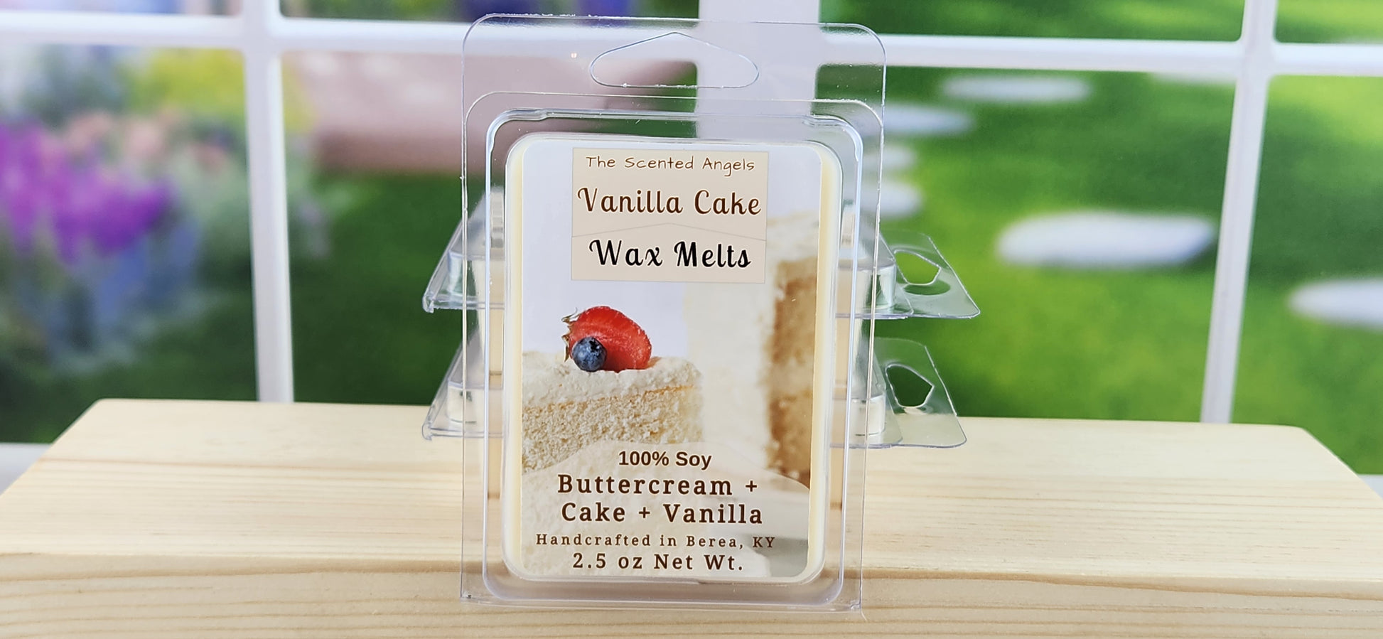 Vanilla cake wax melt packaging with a strawberry on a wooden surface with a garden background.