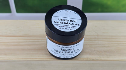 A jar of unscented natural tallow butter with a black lid, labeled with a white and green label, on a wooden surface with a bright background.