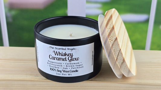 A 5 oz soy wax candle with a label that reads 'Whiskey Caramel Glow', featuring a black matte vessel and a hand-cut pine wood lid, set on a wooden surface.
