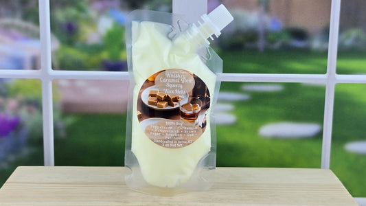 A pouch of Whiskey Caramel Glow 100% Soy Squeezy Wax Melts placed on a wooden surface with a window in the background showing a grassy area.